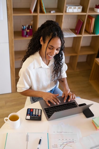 A young Mexican professional using a calculator and a laptop in a modern office setting, analyzing her income and expenses while creating a budget.