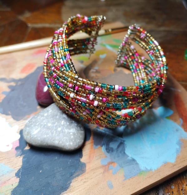 A well-lit showcase of finished artisan jewelry pieces, including colorful beaded earrings, delicate wire-wrapped bracelets with charms, and intricate chaquira necklaces displayed on a velvet surface with soft, diffused lighting to highlight the craftsmanship and details.