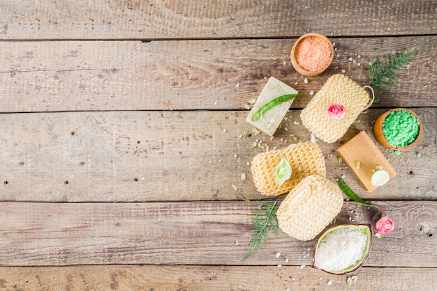 A close-up, overhead shot showing various natural soap ingredients neatly arranged on a wooden surface. The ingredients include dried lavender buds, chamomile flowers, oatmeal, a bottle of olive oil, and a small dish of white clay.