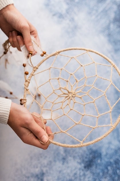 A pair of hands carefully weaving thread around a dreamcatcher hoop, creating the intricate web pattern. The background is blurred, but the focus is on the detail of the weaving process.