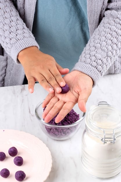 A pair of hands carefully topping a freshly poured beeswax candle with dried lavender buds. The candle is in a small mason jar and the background shows a collection of other candles adorned with different dried herbs and flowers, creating a colorful and artisanal atmosphere.