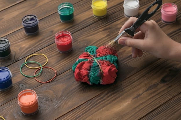 A close-up shot of a crafter's hands carefully arranging various recycled materials, such as colorful paper scraps, buttons, and ribbons, onto a blank greeting card. The hands are holding a small glue bottle, and the background shows a well-organized craft space with containers of pens, scissors, and other tools.