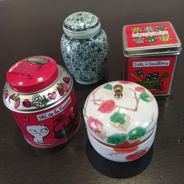 A collection of unique homemade candles in different repurposed containers (old teacups, tin cans, glass jars), arranged on a shelf.