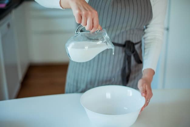 A woman's hands are shown mixing water and fabric softener in a clear glass bowl with a whisk. The bowl sits on a clean kitchen counter with a white tiled backsplash. Sunlight streams in from a nearby window, casting shadows and highlights on the ingredients. The focus is on the mixing process, with the woman's hands and the swirling liquid as the central point.
